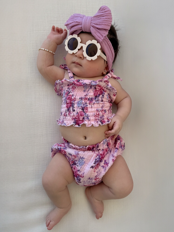 Baby girl in floral outfit with sunglasses and a headband, lying on a light background.