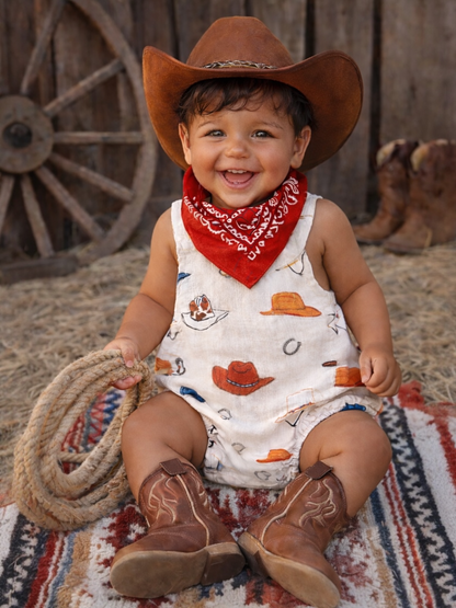 Happy child in cowboy attire with hat and boots, sitting on a colorful blanket in a rustic setting.