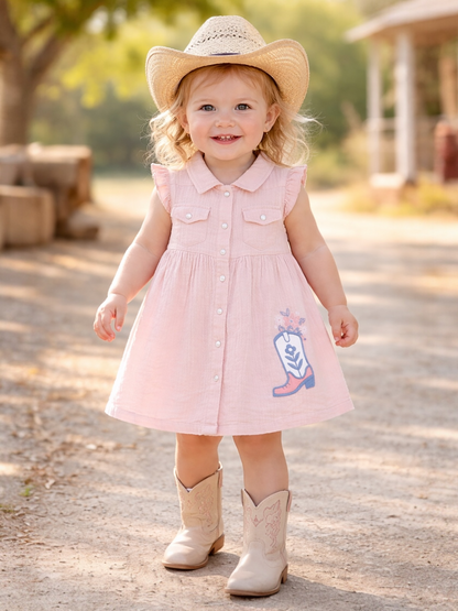 Smiling toddler in a pink dress and cowboy boots stands on a dirt path, wearing a straw hat.