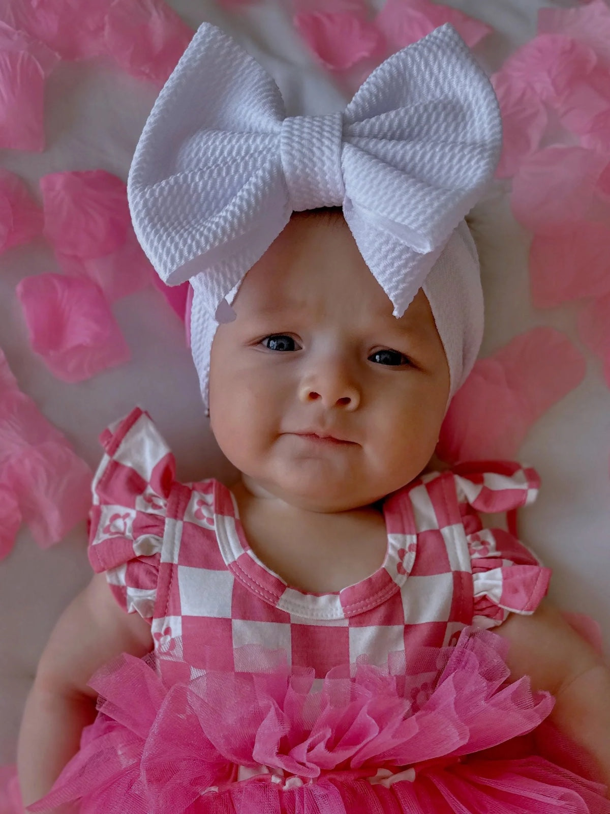 Baby girl with a large white bow, wearing a pink checkered outfit surrounded by pink petals.