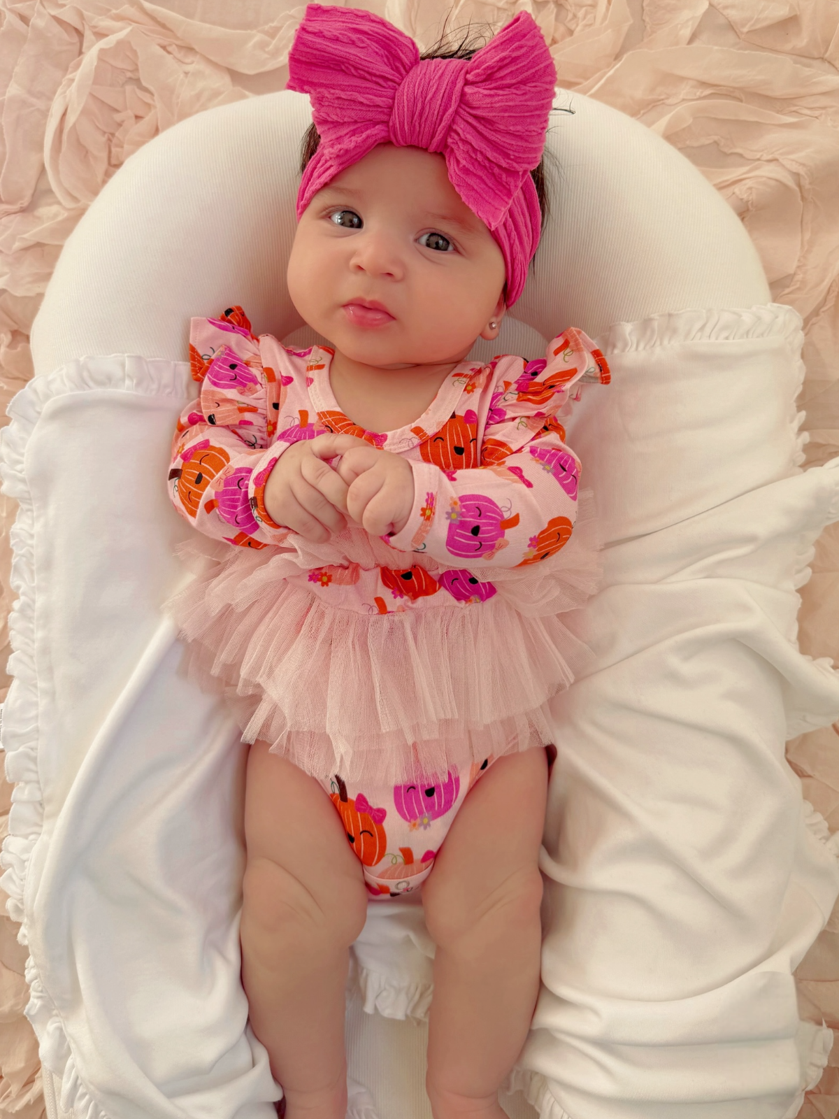 Baby girl in pink outfit with pumpkin print, wearing a large pink bow, sitting on a white plush surface.