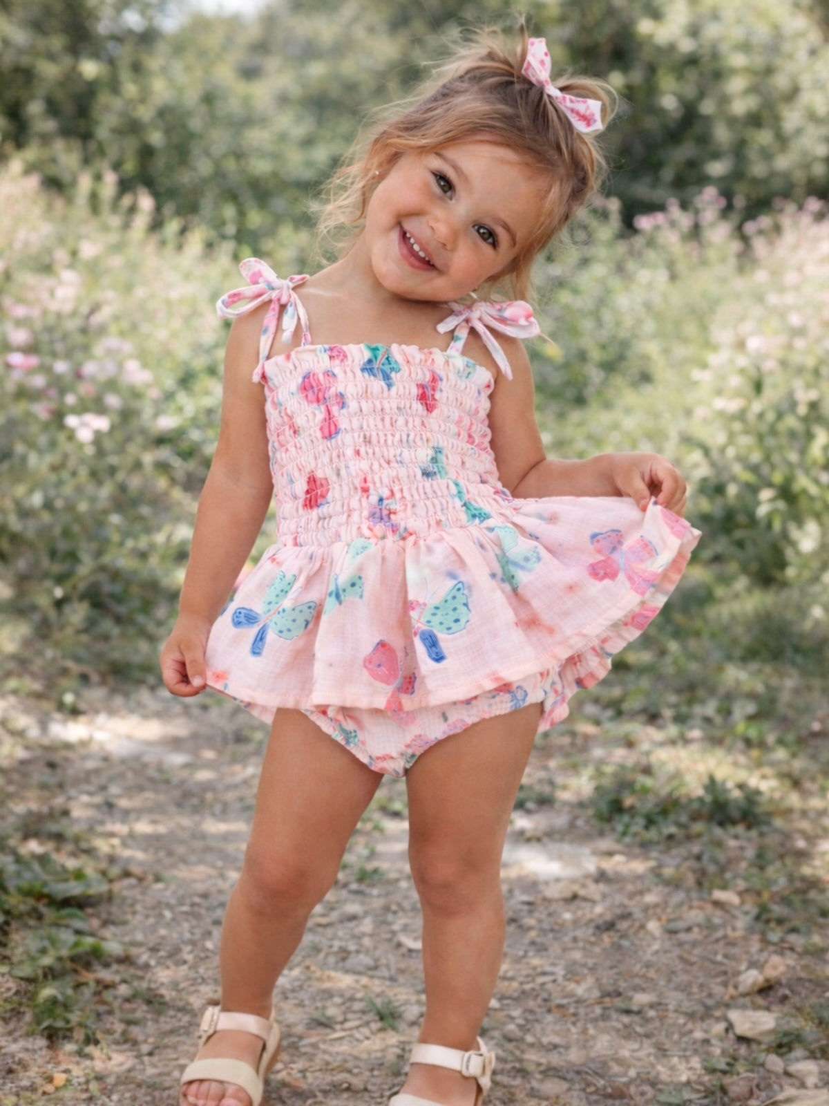 Smiling girl in a pink floral dress stands outdoors, surrounded by greenery and flowers.