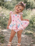 Smiling girl in a pink floral dress stands outdoors, surrounded by greenery and flowers.