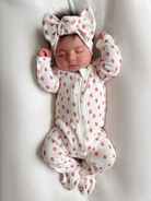 Infant asleep in a strawberry-patterned sleeper and bow headband, laying on a soft white blanket.