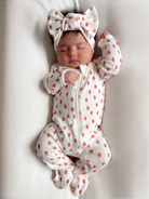 Infant sleeping in a strawberry-patterned onesie and matching bow headband, on a soft, light-colored background.
