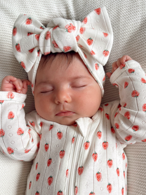 Baby girl sleeping peacefully in a strawberry-patterned outfit and large bow headband on a soft blanket.