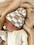 Baby wearing a checkered beanie, peacefully resting on a soft blanket.