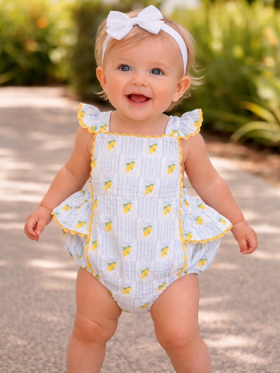 Smiling toddler in a lemon-patterned romper with a white bow, stands on a sunny path surrounded by greenery.