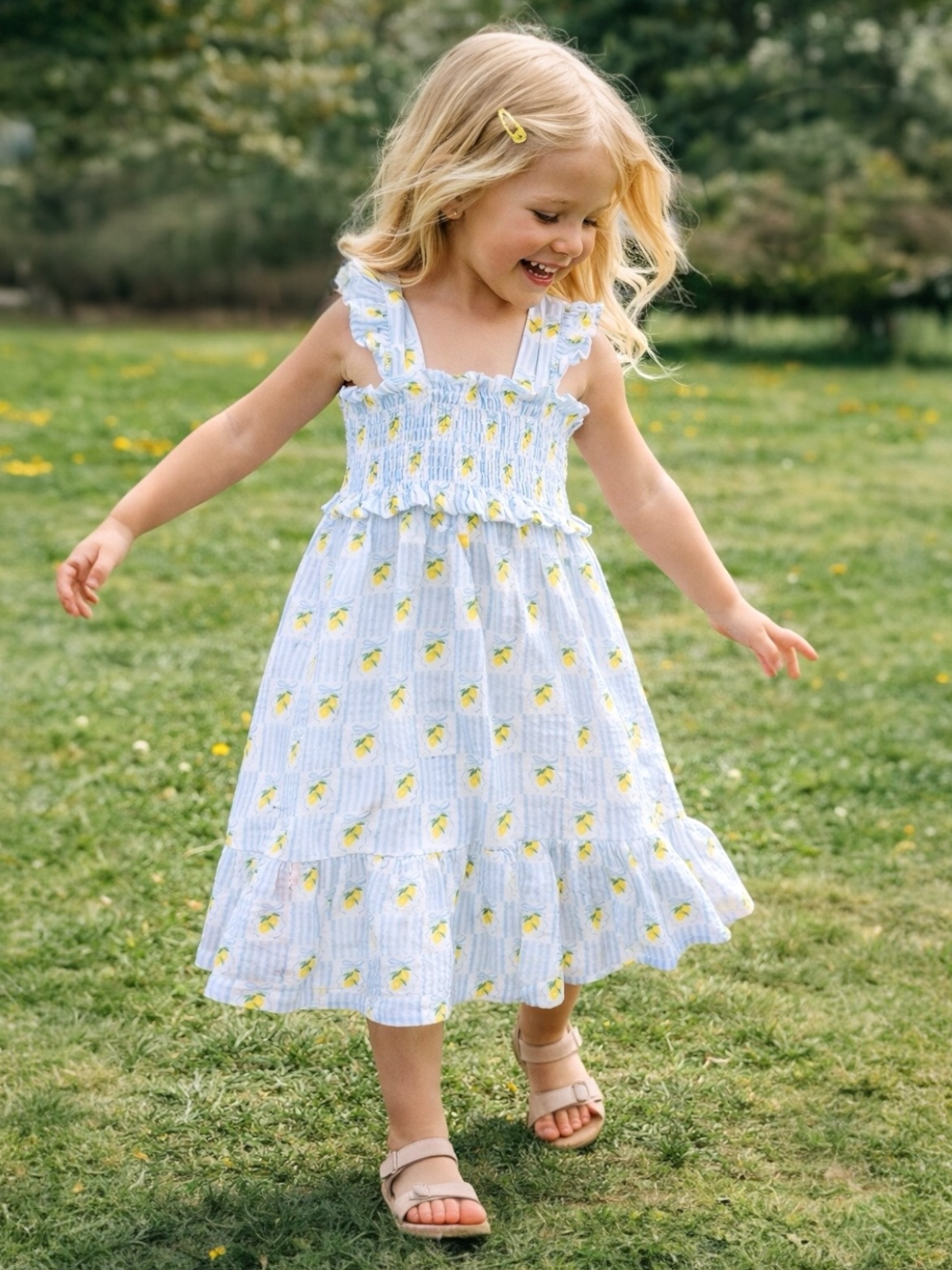 Smiling young girl in a lemon-patterned dress joyfully walks on grass in a sunny outdoor setting.