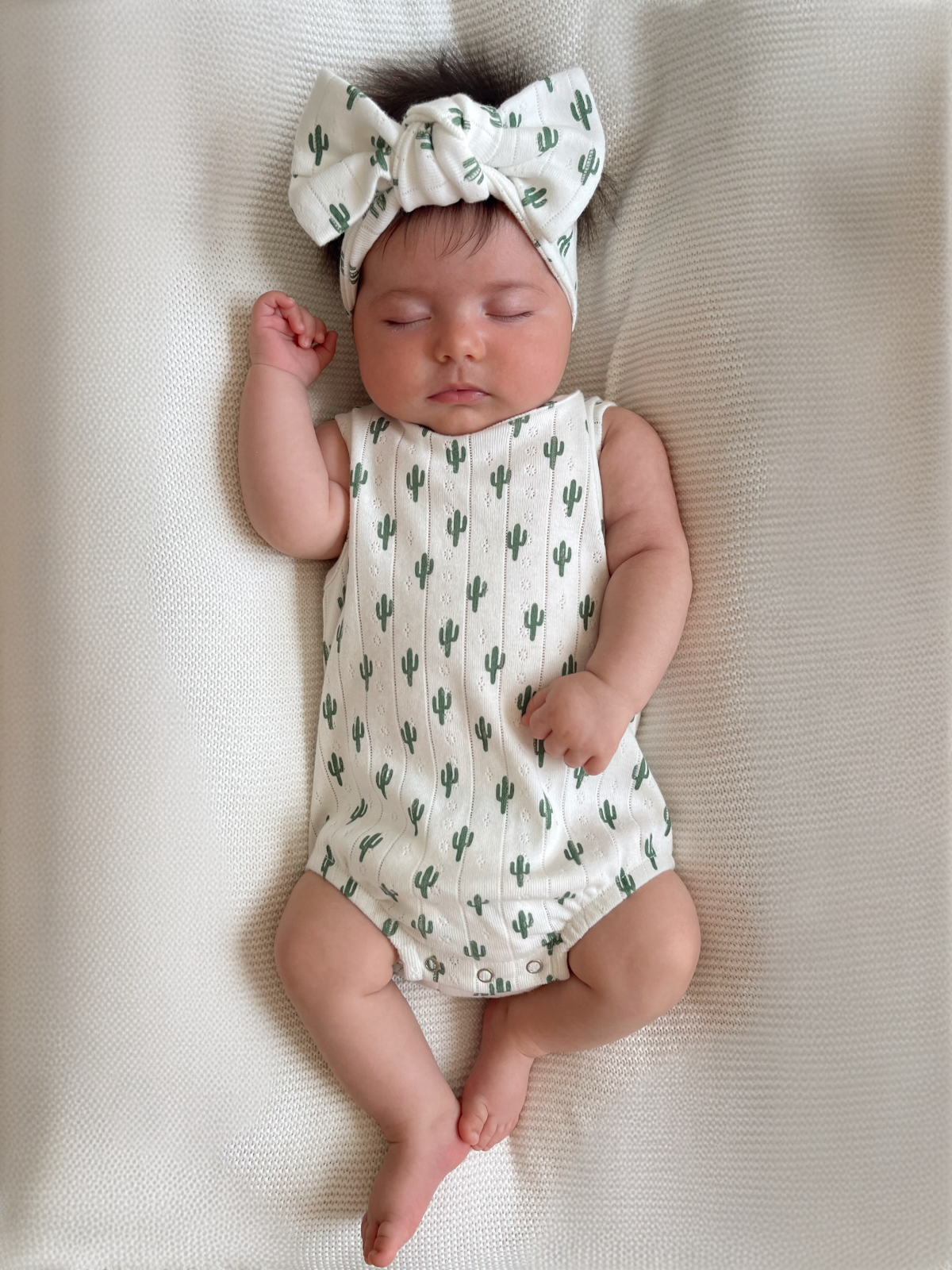 Infant sleeping on a textured blanket, wearing a cactus-patterned onesie and a matching bow headband.