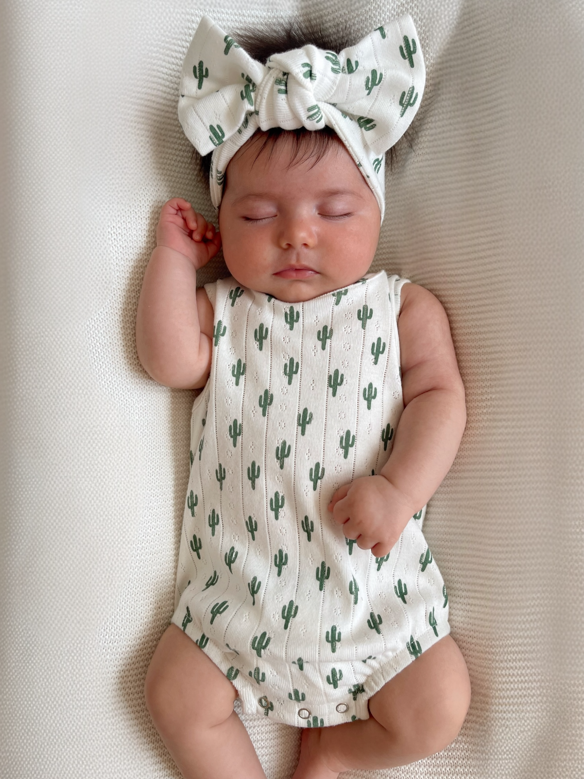 Baby girl sleeping in a cactus-patterned romper and matching bow headband on a textured blanket.