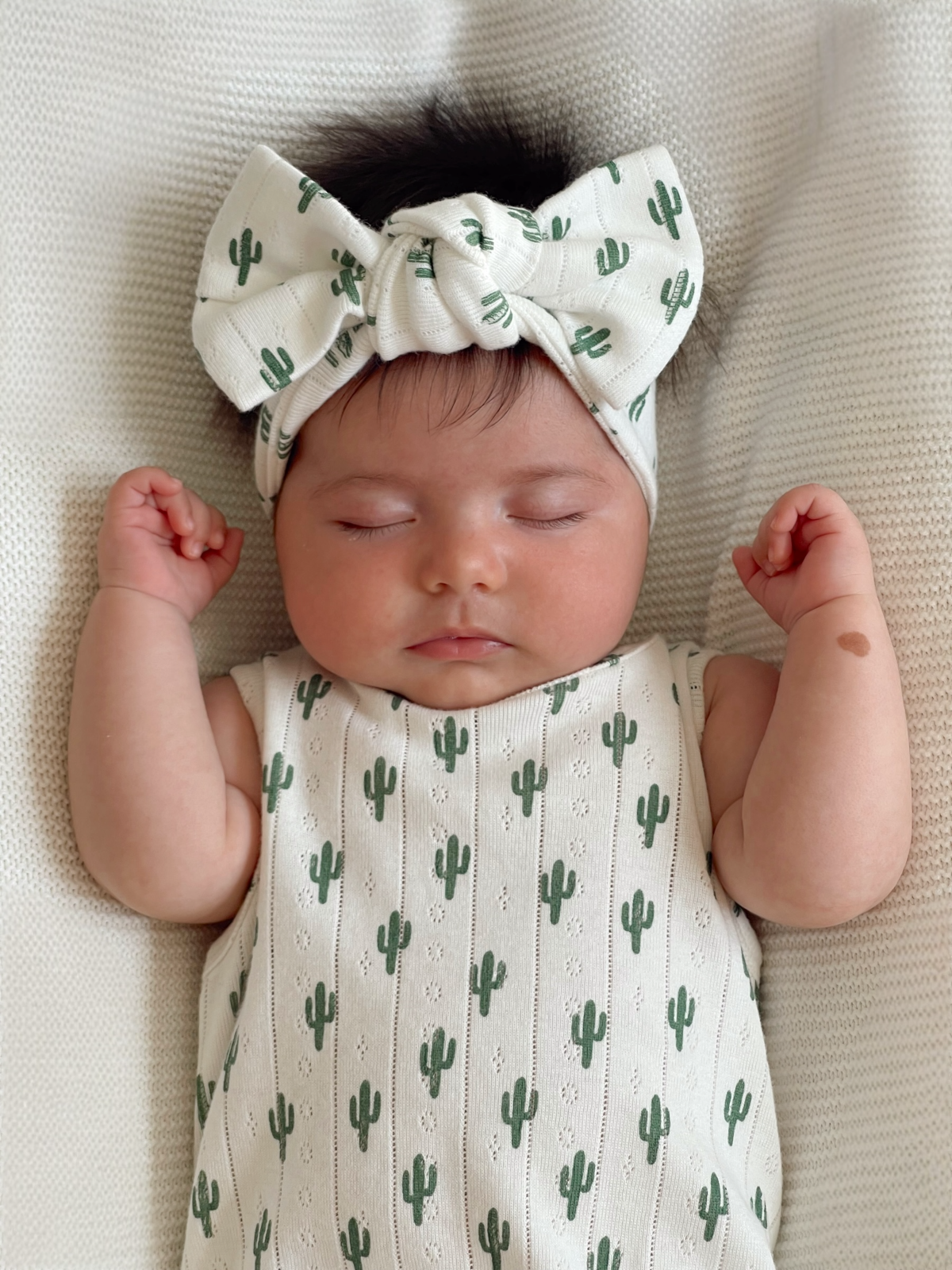 Sleeping baby with a cactus-patterned outfit and a large bow, resting on a textured blanket.