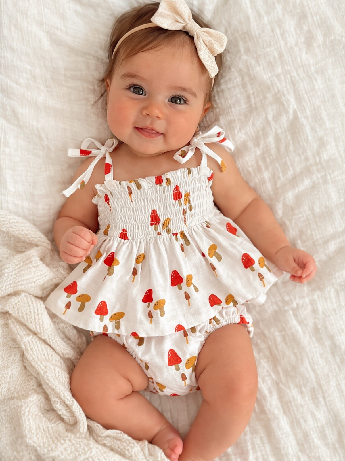 Baby girl in a white mushroom-patterned outfit, smiling on a soft, textured blanket.