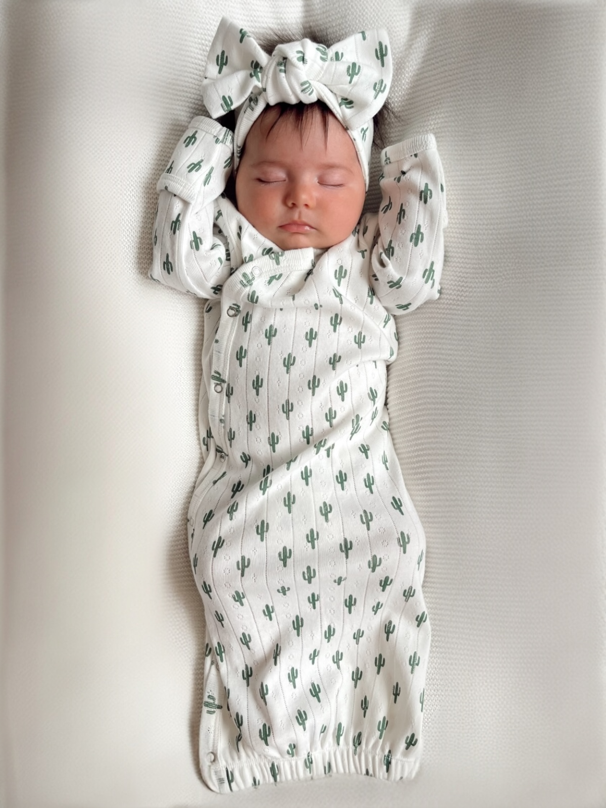 Baby sleeping peacefully in a cactus-patterned swaddle and headband on a soft, textured blanket.