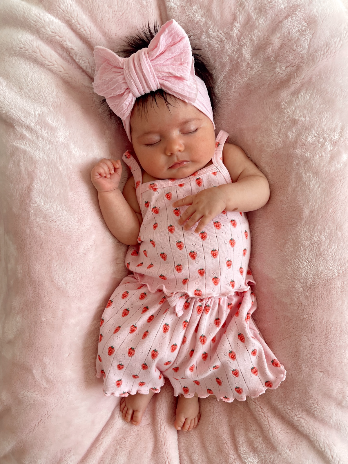 Infant girl asleep on pink blanket, wearing a pink strawberry-patterned outfit and large pink bow headband.