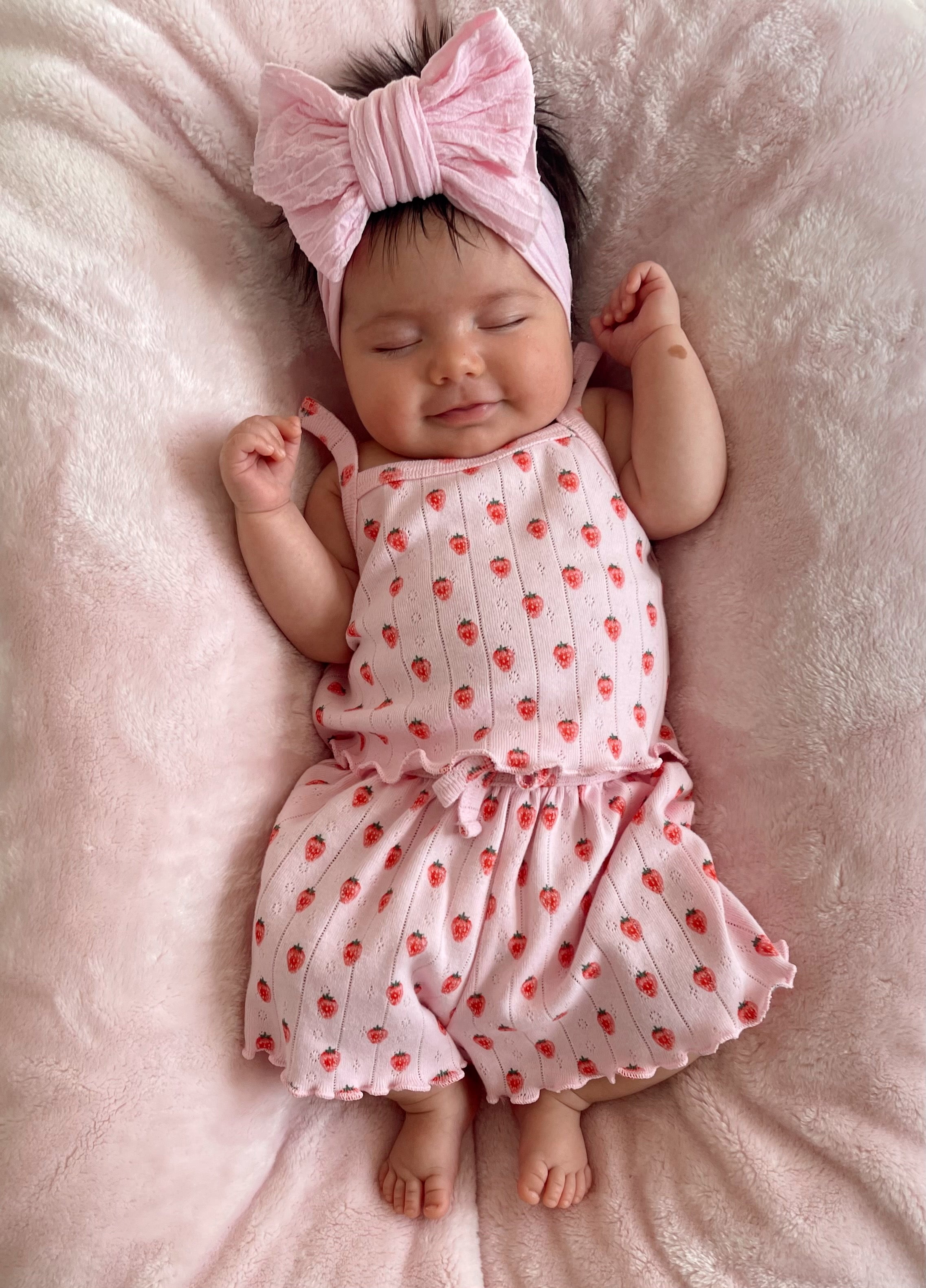 Smiling baby in a pink strawberry-patterned outfit with a large bow headband, lying on a soft pink blanket.
