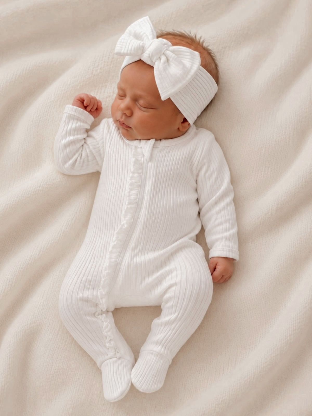 Newborn baby girl sleeping peacefully in a white ribbed onesie with a large bow headband on a soft blanket.