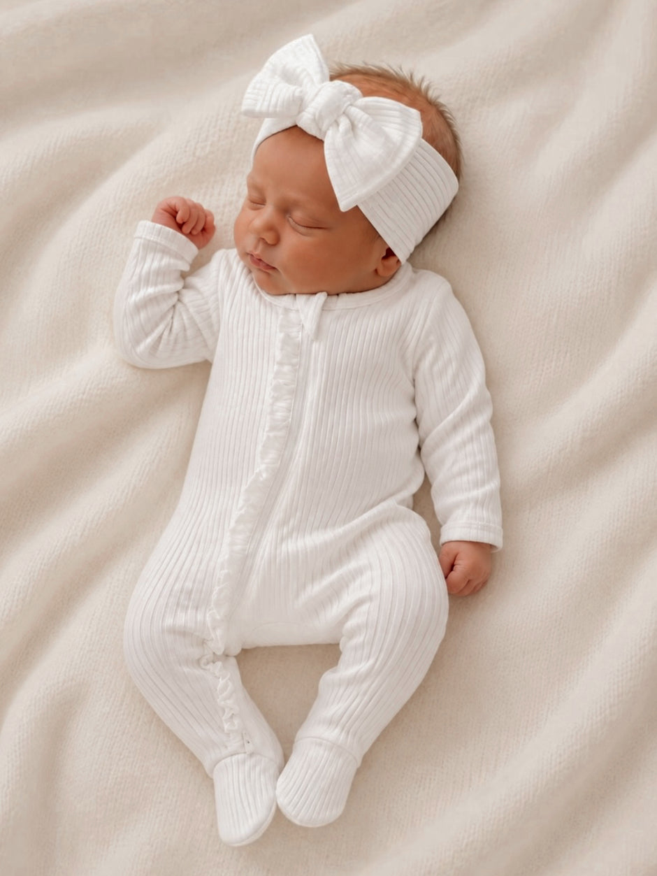 Newborn baby girl sleeping peacefully in a white ribbed onesie with a large bow headband on a soft blanket.