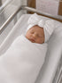 Newborn baby girl swaddled in white fabric, wearing a white headband with a large bow, peacefully sleeping.