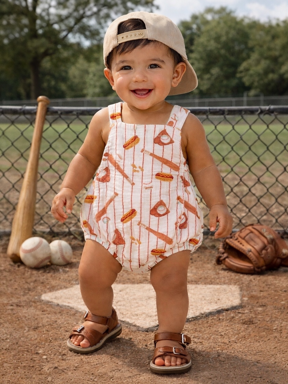 Smiling toddler in a baseball-themed outfit stands on a dirt field, wearing a cap and sandals.