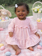 Smiling baby girl in a pink dress, surrounded by pastel Easter eggs and a bunny toy on a blanket outdoors.