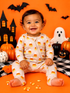 Smiling baby in candy corn pajamas surrounded by Halloween decorations on an orange background.