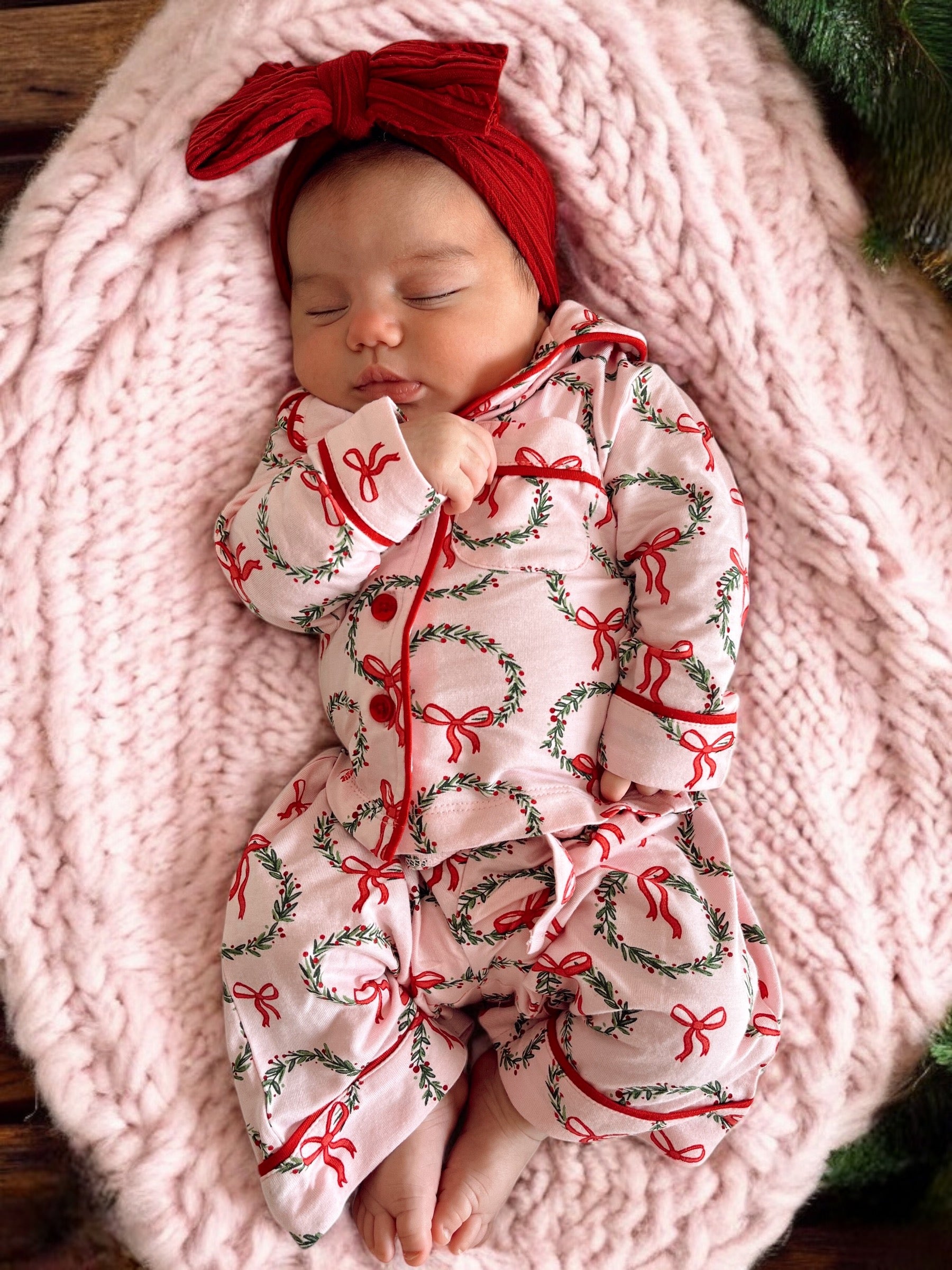 Baby girl in a pink holiday onesie with wreaths, resting on a soft pink blanket, wearing a red headband.
