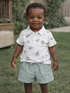 Smiling toddler in a turtle-patterned shirt and green shorts, standing on grass with a wooden backdrop.