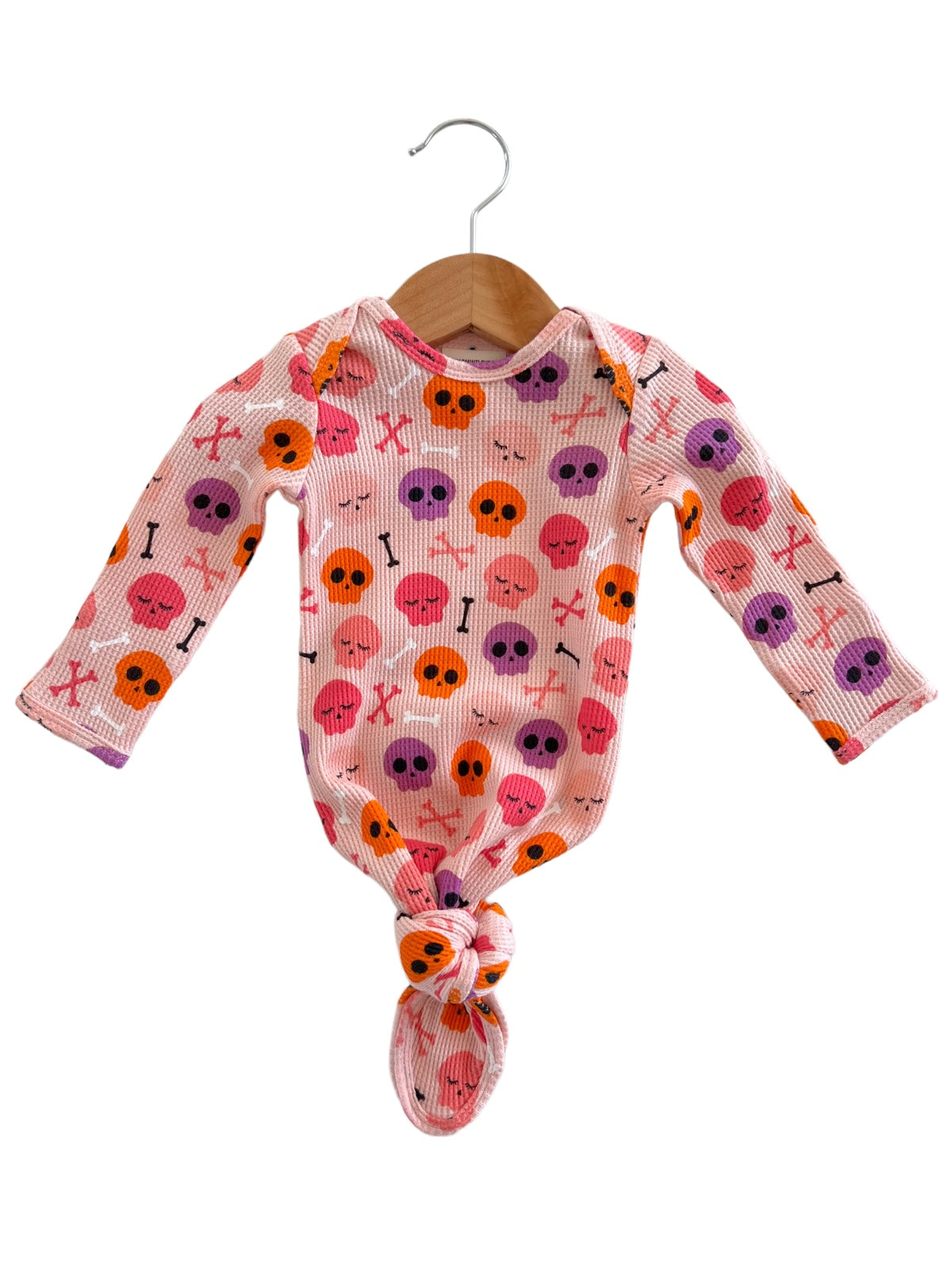 Pink baby onesie with colorful skull and crossbone pattern, tied at the bottom, hanging on a wooden hanger.