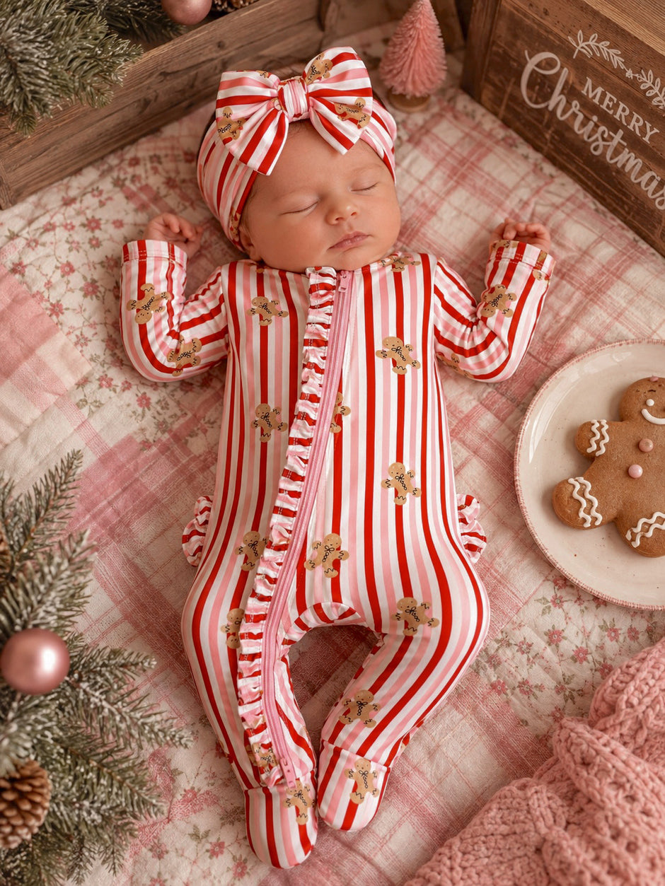 Baby girl sleeping in a festive striped onesie with gingerbread men, wearing a matching headband, surrounded by holiday decor.