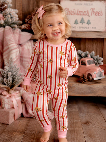 Smiling toddler in striped pajamas with gingerbread prints, surrounded by holiday decor and gifts.