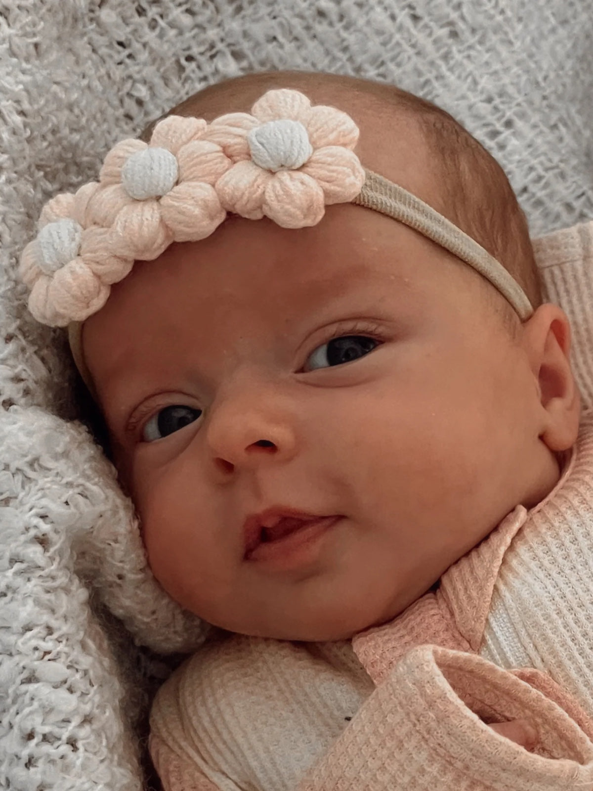 Infant girl with a flower headband gazing into the camera, wrapped in a cozy blanket.