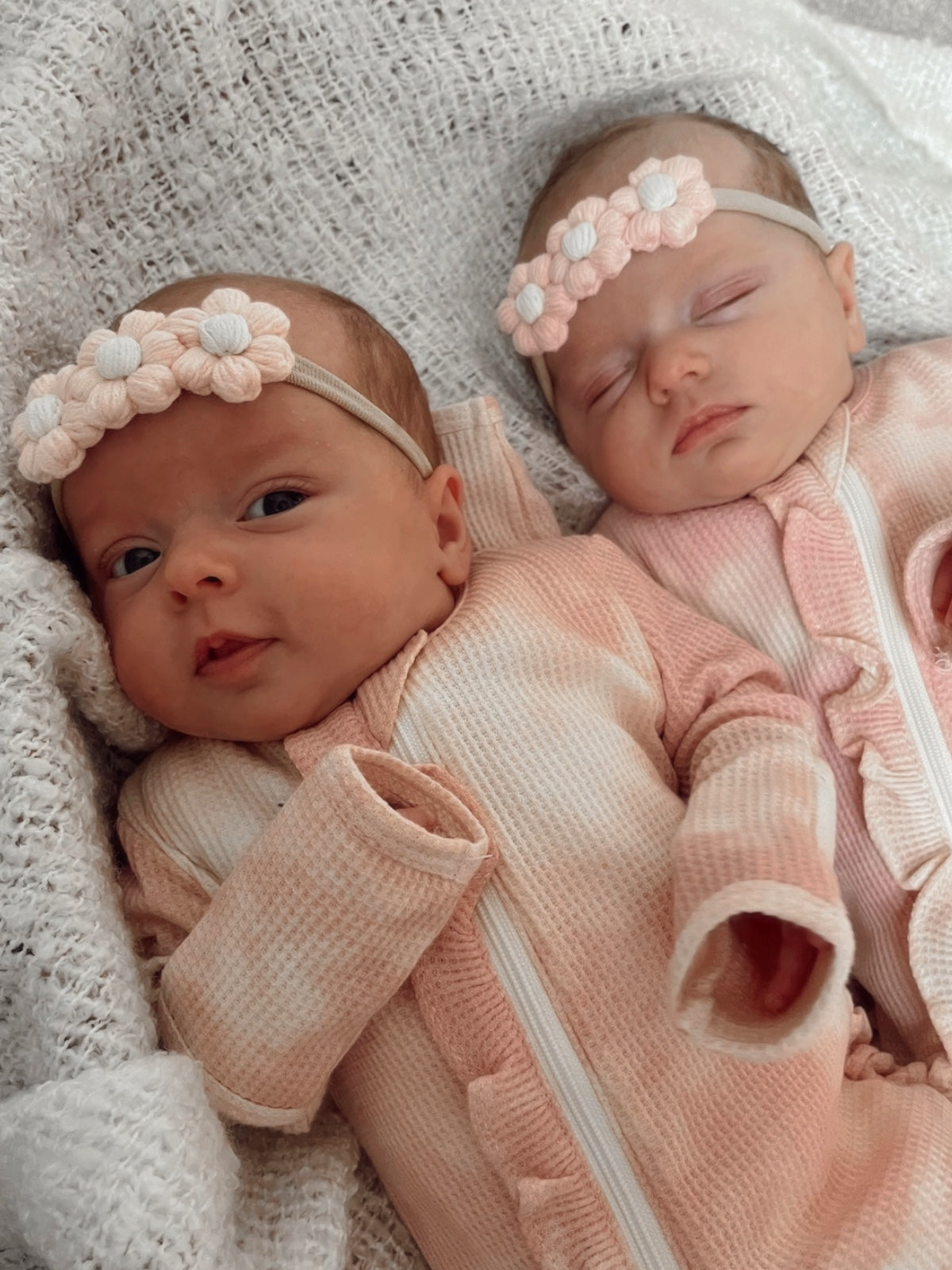 Two adorable newborns in pink outfits with floral headbands, snuggled together on a cozy blanket.