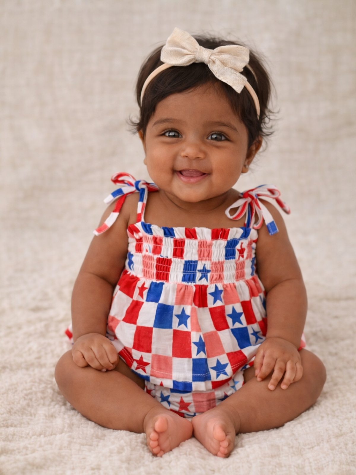Smiling baby girl in red, white, and blue checkered outfit with a bow, sitting on a soft surface.