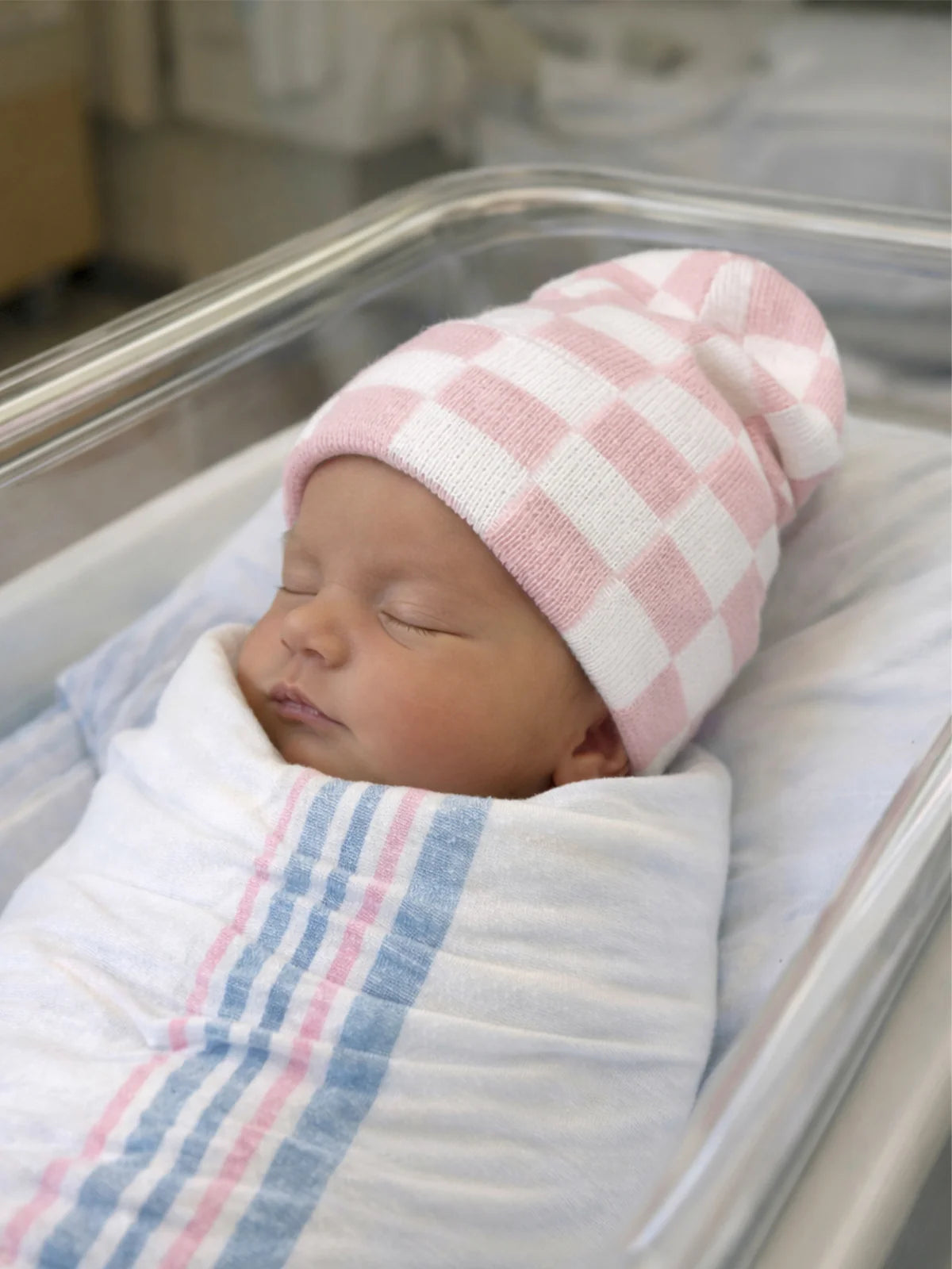 Newborn baby wrapped in a blanket, wearing a pink and white checkered hat, peacefully asleep in a hospital bassinet.