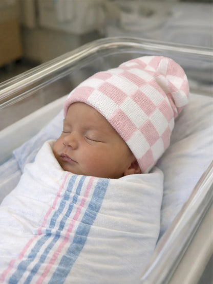 Newborn baby wrapped in a blanket, wearing a pink and white checkered hat, peacefully asleep in a hospital bassinet.