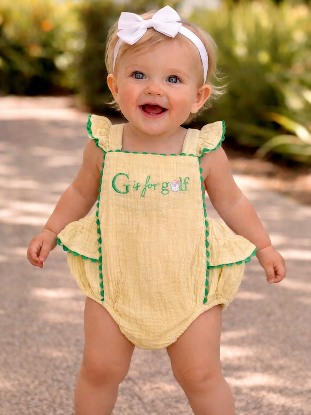 Smiling baby in a yellow romper with "G is for golf," wearing a white bow, standing outdoors with greenery.