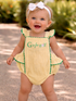 Smiling baby in a yellow romper with "G is for golf," wearing a white bow, standing outdoors with greenery.