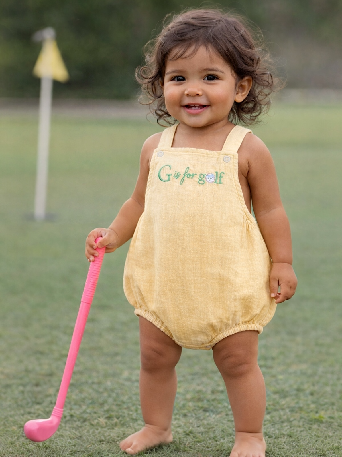 Smiling toddler in yellow outfit holding a pink golf club on a grassy field, sunny day.