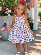 Smiling girl in a patriotic dress holding a small American flag, surrounded by colorful flowers.