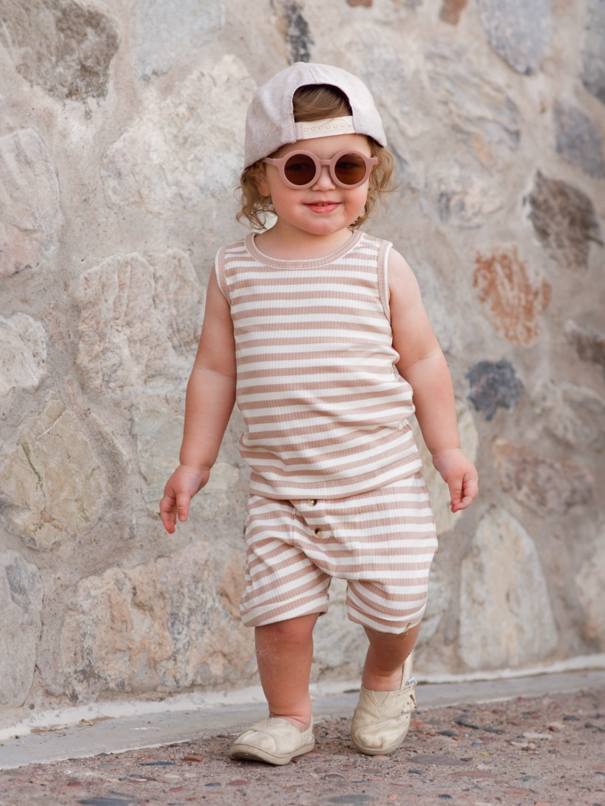 Smiling toddler in striped outfit, cap, and sunglasses, walking on a stone path.