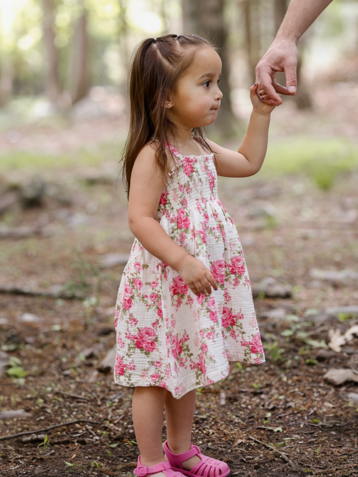 Young girl in a floral dress holds an adult's hand in a wooded area.