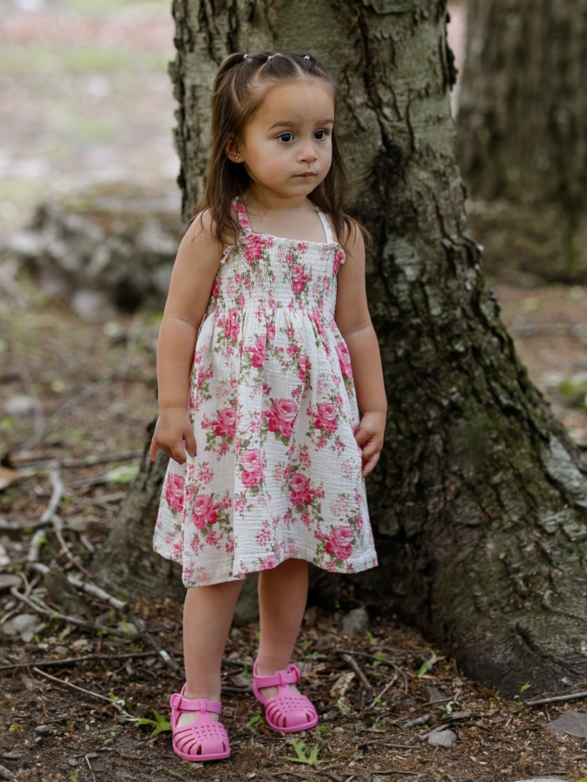 Young girl in a floral dress stands beside a tree, wearing pink sandals in a natural outdoor setting.