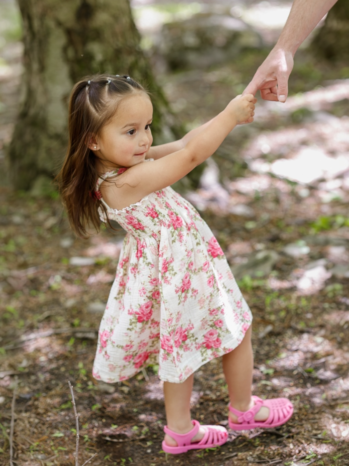 Young girl in a floral dress holds hands with an adult in a sunlit forest setting.