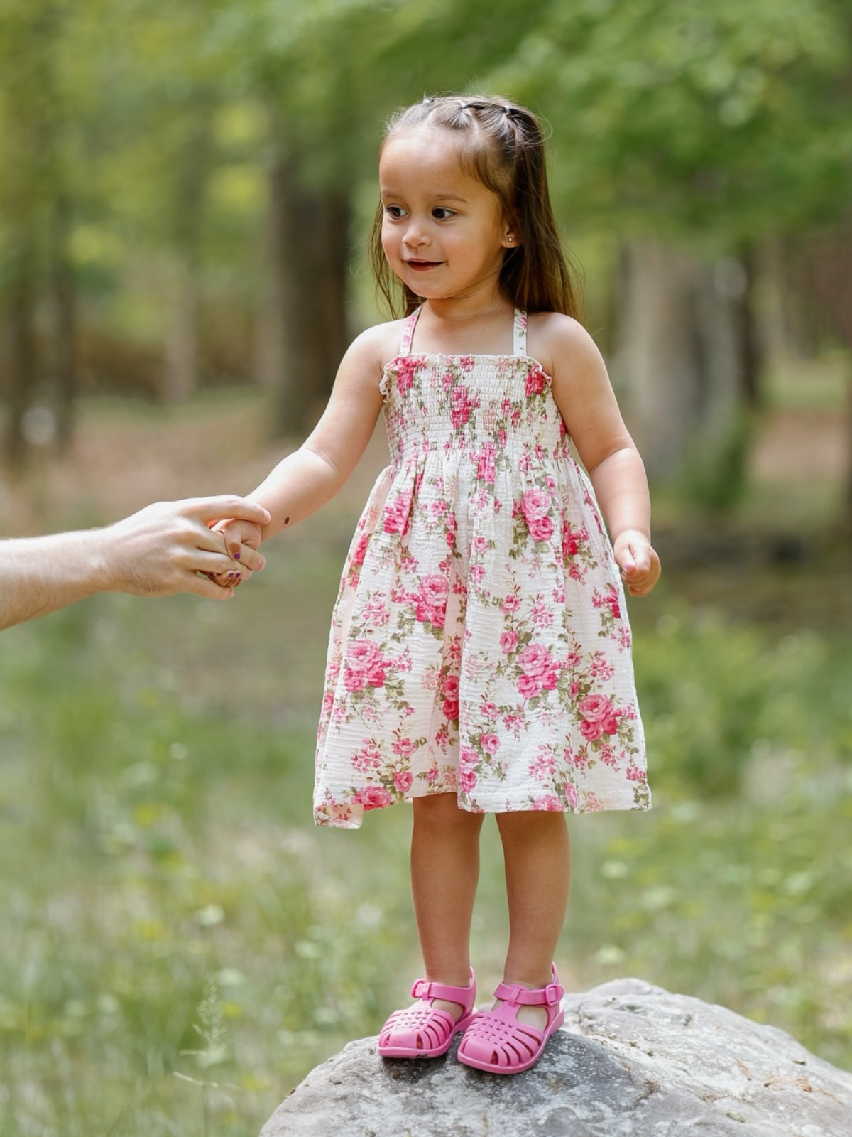 Little girl in a floral dress stands on a rock, holding hands with an adult in a green forest setting.