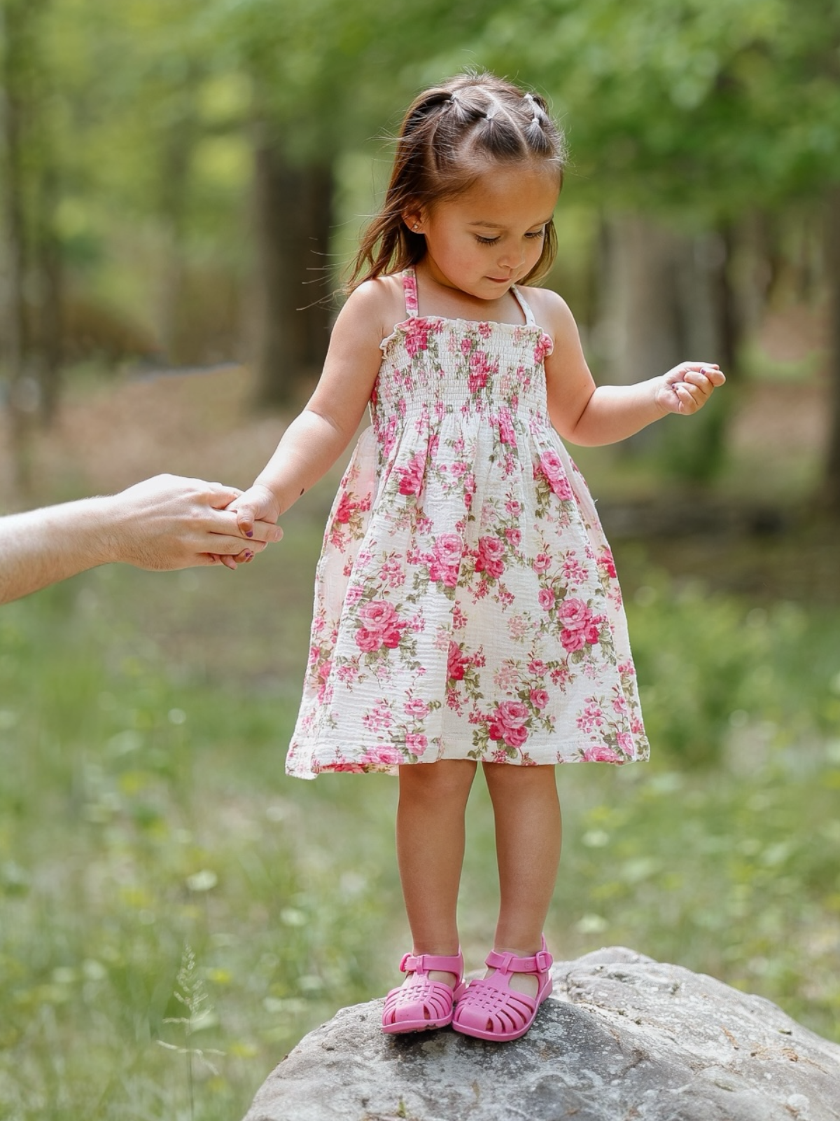 Young girl in a floral dress stands on a rock, holding an adult's hand in a lush, green outdoor setting.