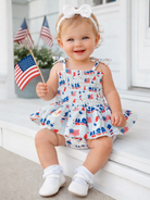 Smiling toddler in a patriotic outfit, holding an American flag, sitting on steps with flags in the background.