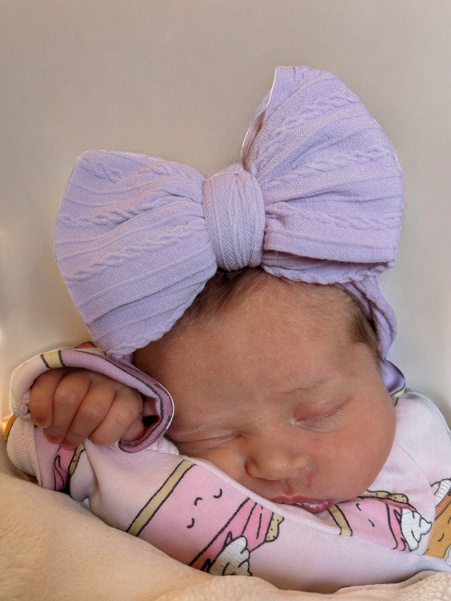 Newborn girl sleeping with a large lavender bow and pink ice cream-themed outfit, gently holding her hand.