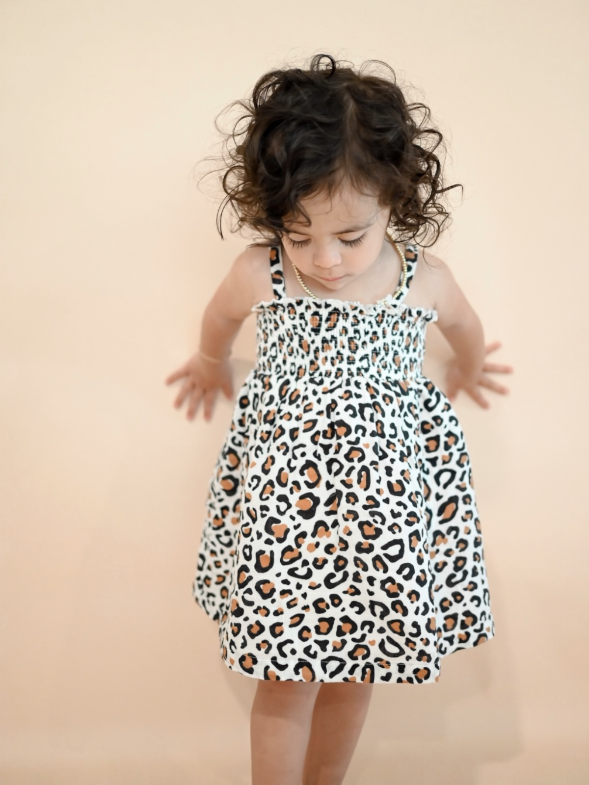 Child with curly hair wearing a leopard print dress, standing against a soft peach background.