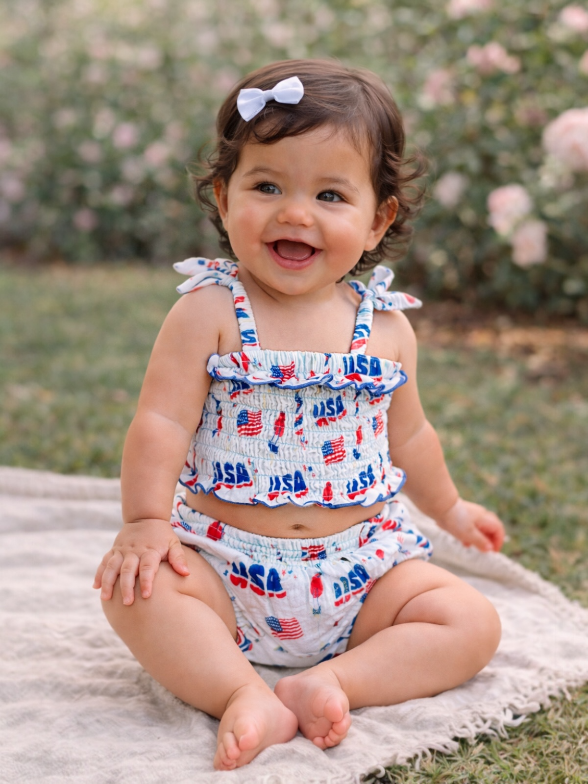 Smiling baby in patriotic outfit sitting on a blanket with roses in the background.